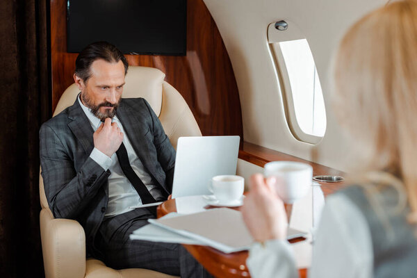 Selective focus of businessman using laptop near businesswoman drinking coffee in plane 