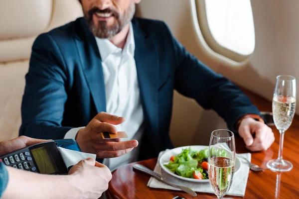 笑顔のビジネスマンの近くにターミナルを保持する男の作物ビュークレジットカード サラダ シャンパンでテーブルの上に平面 — ストック写真