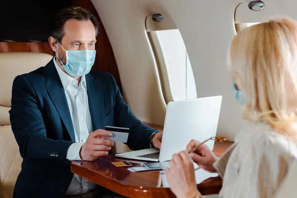 商人佩戴医疗面罩 持有信用卡 在飞机上使用手提电脑的选择性焦点 — 图库照片