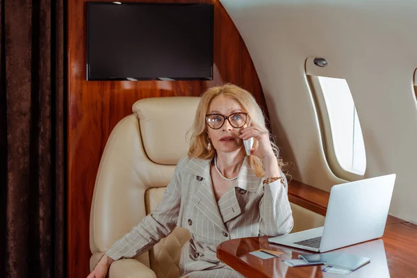 飛行機のテーブルの上でクレジットカードやノートパソコンの近くのスマートフォンで話しているビジネスマンの女性 — ストック写真