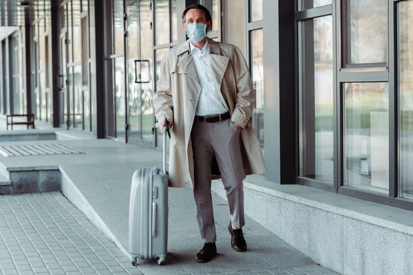 一个戴医疗面具的英俊商人拿着手提箱靠近市区街上的大楼 — 图库照片