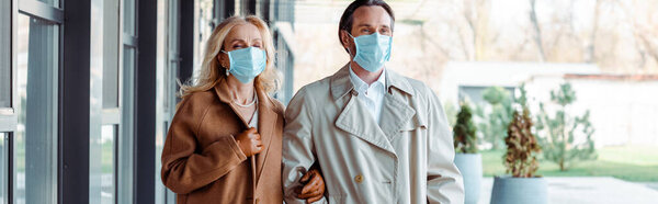 Panoramic shot of business couple in medical masks looking away near building on urban street 