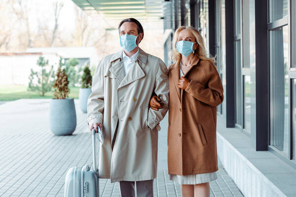 Зрелая деловая пара в медицинских масках, стоящая возле чемодана на городской улице
 