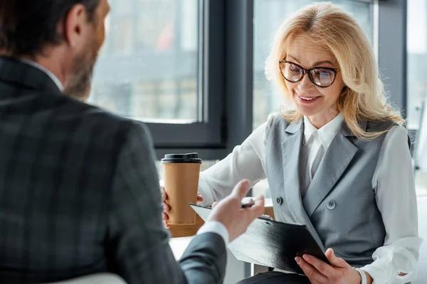 Selektywne Skupienie Uśmiechniętej Bizneswoman Kawą Iść Patrząc Schowek Pobliżu Biznesmena — Zdjęcie stockowe