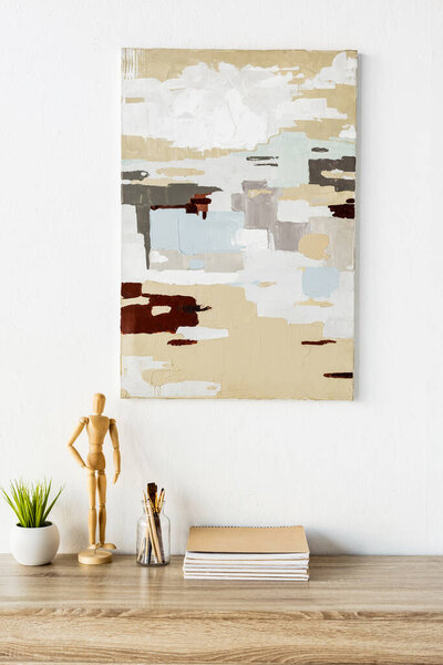 painting on wall near table with wooden mannequin, plant, notebooks, paintbrushes and pencils 