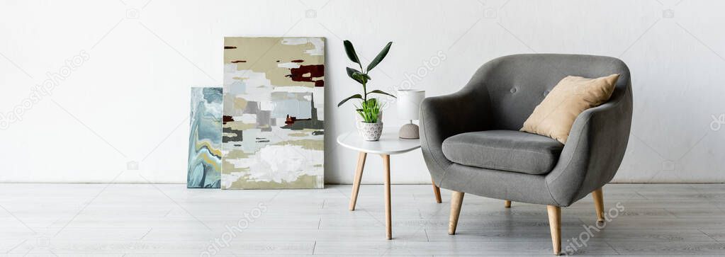 panoramic crop of comfortable armchair near coffee table with green plants, lamp and paintings in living room 