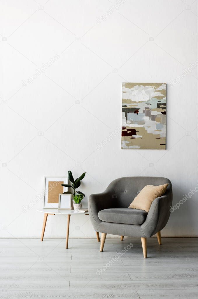 grey armchair near coffee table with frames and green plants near painting on wall in modern living room 