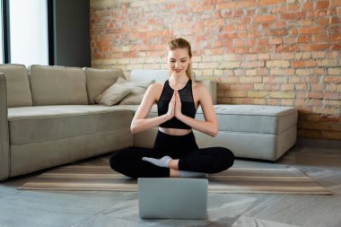 Dua eden elleri olan mutlu sporcu kadın oturma odasında dizüstü bilgisayarında yoga egzersizi izliyor. 