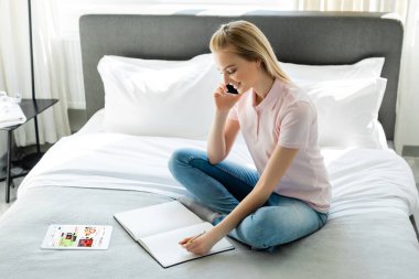 KYIV, UKRAINE - 8 Nisan 2020: mutlu kadın defterine bakıyor ve yatak odasında ebay uygulaması olan dijital tabletin yanında akıllı telefonla konuşuyor 