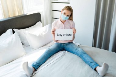 woman in medical mask holding paper with stay at home lettering while sitting on bed  clipart