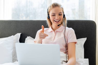 Yatak odasındaki bilgisayarı işaret eden kulaklıklı neşeli serbest çalışanın seçici odağı 
