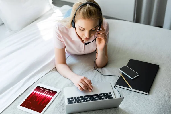 戴耳机的年轻自由职业者 在数码平板电脑旁使用笔记本电脑 附带交易课程 在床上使用带有空白屏幕的智能手机 — 图库照片