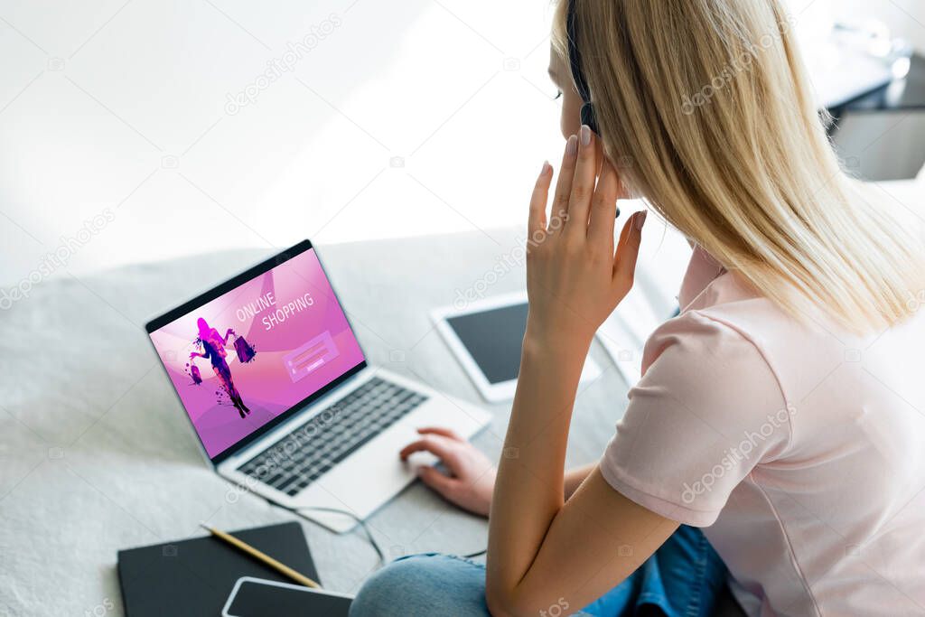 selective focus of freelancer touching headset near laptop with online shopping website and digital tablet with blank screen on bed