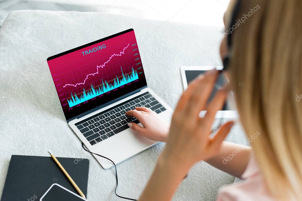 selective focus of freelancer touching headset near laptop with trading and digital tablet with blank screen on bed