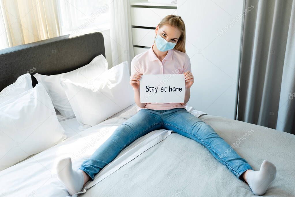 woman in medical mask holding paper with stay at home lettering while sitting on bed 