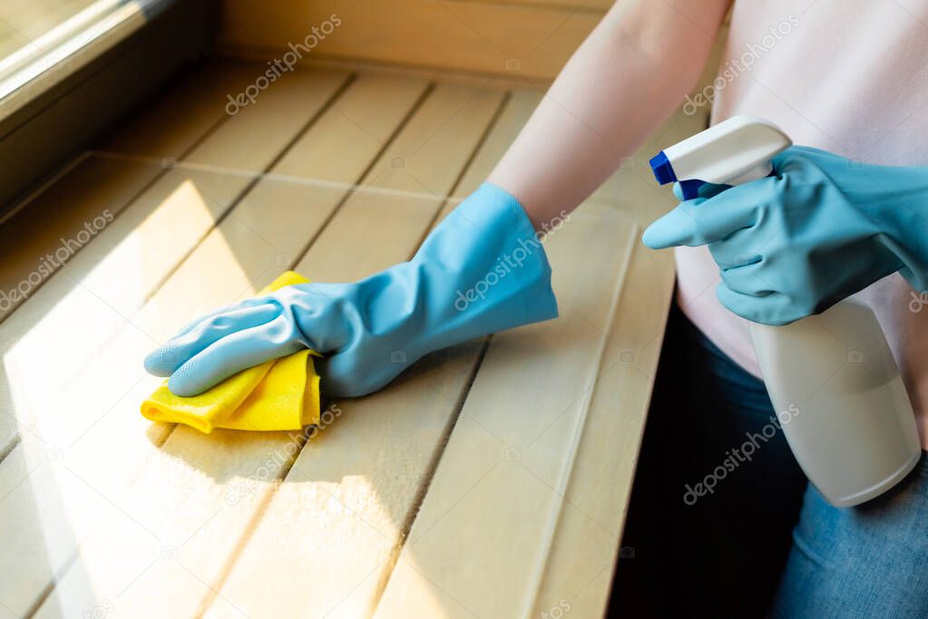 cropped view of girl in rubber gloves holding spray bottle and rag near wooden surface 