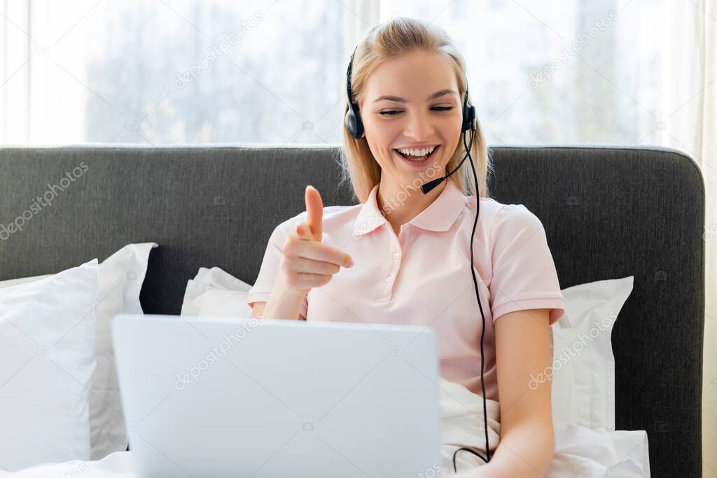 selective focus of cheerful freelancer in headset pointing with finger at laptop in bedroom 