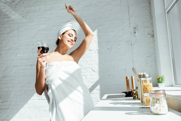 Smiling woman in towels holding glass of wine near kitchen worktop 