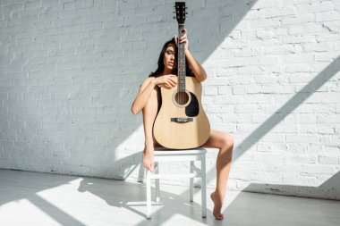 Oturma odasında sandalyede otururken akustik gitar tutan güzel bir kadın.