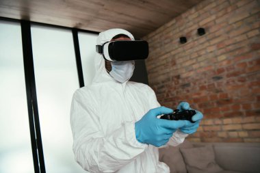KYIV, UKRAINE - 11 Nisan 2020: Tehlikeli madde giysili adam, tıbbi maske, lateks eldivenler ve oturma odasında video oyunu oynayan sanal gerçeklik kulaklığı 