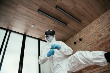 Tehlikeli madde giysisi giymiş bir adamın düşük açılı görüntüsü, tıbbi maske, lateks eldivenler ve oturma odasındaki sanal gerçeklik kulaklığı. 
