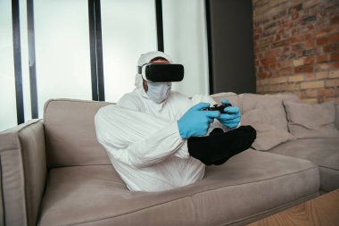 KYIV, UKRAINE - 11 Nisan 2020: Tehlikeli madde giysili adam, tıbbi maske, lateks eldiven ve kanepede video oyunu oynayan sanal gerçeklik kulaklığı
