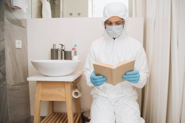 Tehlikeli madde giysili, tıbbi maskeli, lateks eldivenli ve gözlüklü bir adam banyoda kitap okuyor. 