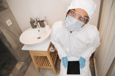 Tehlikeli madde giysili, tıbbi maskeli, lateks eldivenli ve gözlüklü bir adam banyoda boş ekranlı dijital tablet tutuyor. 