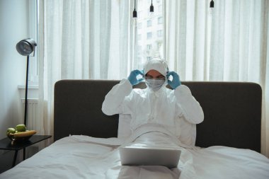 Tehlikeli madde giysisi, tıbbi maske, lateks eldivenler ve gözlükler takan kulaklıklara dokunan ve yatak odasında dizüstü bilgisayar kullanan serbest çalışan biri. 