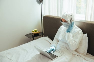Tehlikeli madde giysisi, tıbbi maske, lateks eldivenler ve gözlükler takan kulaklıklara dokunan ve yatakta dizüstü bilgisayar kullanan serbest çalışan biri.