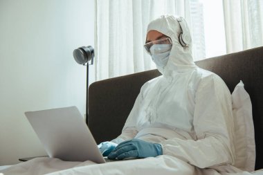 Tehlikeli madde giysisi, kulaklık, tıbbi maske, lateks eldiven ve gözlük giyen yatak odasında dizüstü bilgisayar kullanan serbest çalışan biri. 