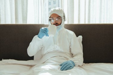 Tehlikeli madde giysisi, kulaklık, tıbbi maske, lateks eldiven ve gözlük takan adam yatak odasında. 