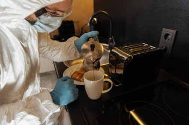 selective focus of man in personal protective equipment pouring coffee in cup near toast bread and toaster in kitchen  clipart