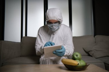 Kişisel koruyucu ekipman ve tıbbi maske giyen bir adam oturma odasında dijital tablet kullanıyor. 