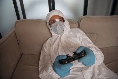 KYIV, UKRAINE - 11 Nisan 2020: oturma odasında video oyunu oynayan kişisel koruyucu ekipman ve tıbbi maske giyen adam 