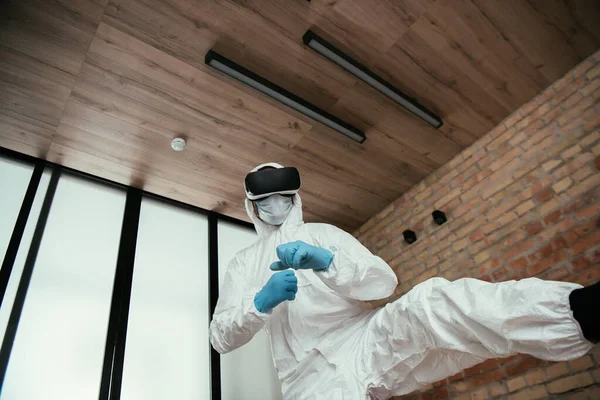 ハズマットスーツの男の低角度ビュー 医療用マスク ラテックス手袋 リビングルームでの仮想現実ヘッドセットゲーム — ストック写真