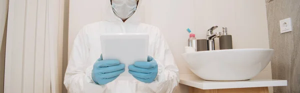 Tehlikeli madde giysisi giymiş bir adamın panoramik görüntüsü, tıbbi maske ve lateks eldivenler banyoda dijital tablet kullanıyor. 