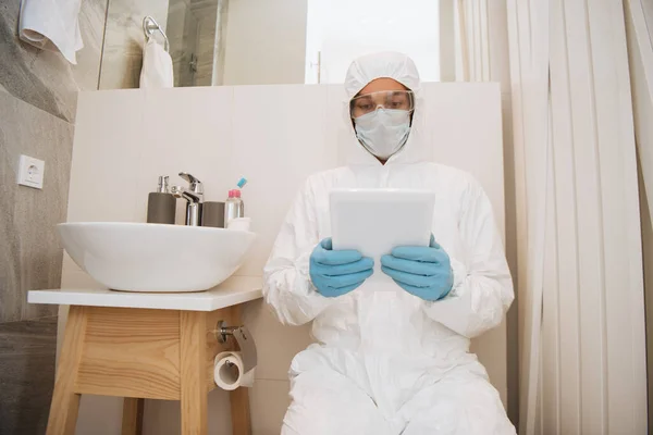在浴室使用数码平板电脑 穿着危险垫套装 护目镜 医疗面罩和乳胶手套的男子 — 图库照片