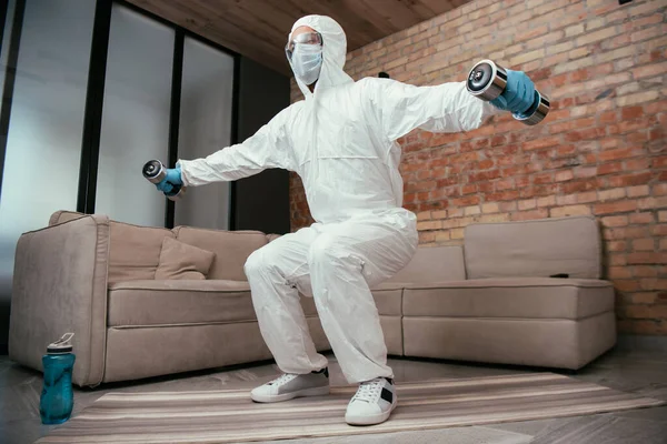sportive man in hazmat suit, medical mask and goggles doing squat exercise with dumbbells near sports bottle and sofa in living room