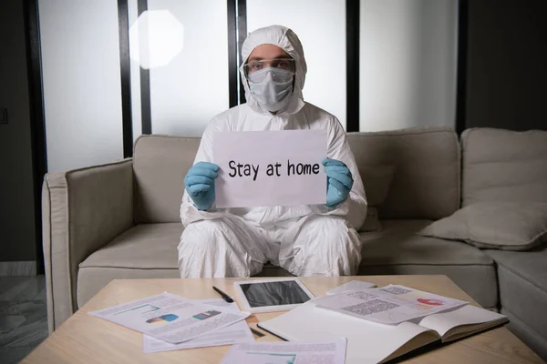 穿着个人防护装备和乳胶手套的男人拿着纸 待在家里 在带空白屏幕的数码平板电脑旁刻有字母 — 图库照片