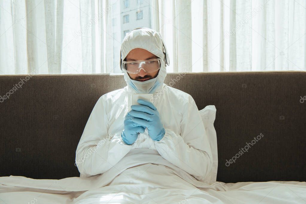 man in hazmat suit, headphones, medical mask, latex gloves and goggles holding cup with drink in bed