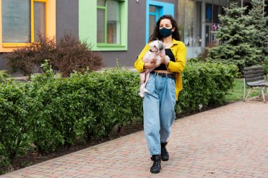 Tıbbi maskeli genç bir kadın şehirde köpekle yürüyor.