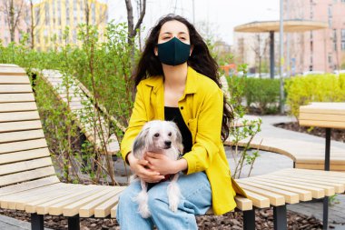 Siyah tıbbi maskeli güzel bir kadın parktaki bankta Çin armalı bir köpekle oturuyor. 