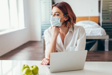 Medikal maskeli düşünceli serbest çalışan evde dizüstü bilgisayarla çalışan kendini izole eden elmalarla. 