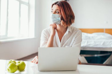 Medikal maskeli kadın evde karantinada elmalarla dizüstü bilgisayarda çalışıyor.