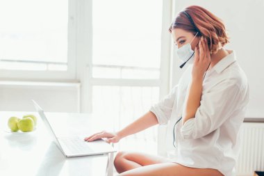 Medikal maskeli mutlu serbest çalışan kulaklık ve dizüstü bilgisayar kullanarak evden kendini izole ediyor.