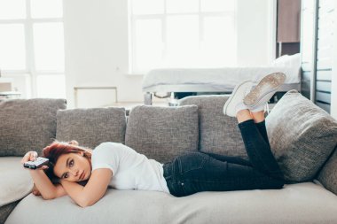 Uzaktan kumandalı kız kendini tecrit sırasında kanepede yatarken televizyon izliyor.
