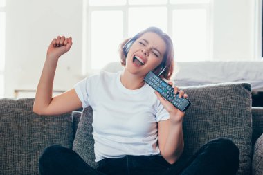 excited girl in headphones singing with remote controller at home during self isolation clipart