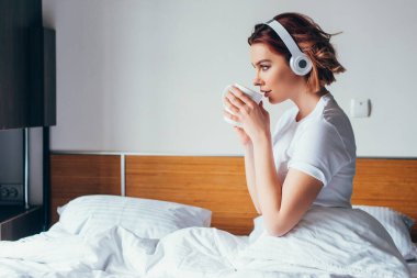 Karantina sırasında yatakta kulaklıkla kahve içip müzik dinleyen çekici genç bir kadın.
