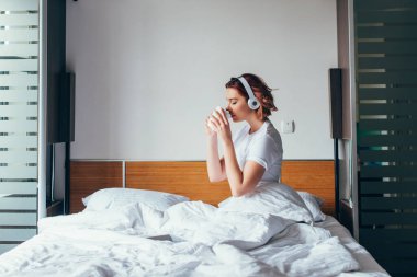 Kendini izole ederken yatakta kahve içerken kulaklıkla müzik dinleyen çekici kız.